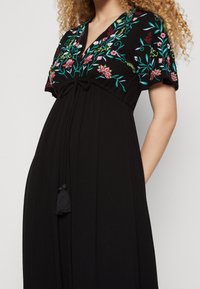 Schwarzes Kleid mit floralen Stickereien in Pink und Grün. Es hat einen V-Ausschnitt, kurze Ärmel und einen Kordelzug an der Taille mit einem Quasten.