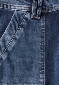 Denimstoff in einem mittleren Blauton, mit auffälligen Nähten, einer glatten Textur und sichtbaren Taschen. Nahaufnahme der Webart des Materials.
