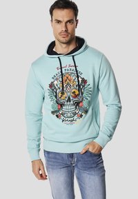 Jasnoniebieska bluza z kapturem z grafiką czaszki i kwiatowymi akcentami; posiada kaptur wyłożony czarną tkaniną, sznurki do regulacji oraz ściągacze na mankietach i u dołu.