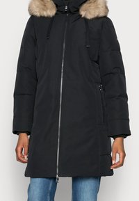 Sort knælængde vinterfrakke med hætte trimmet med pels, frontlynlås, lange ærmer og lynlåslommer på siden, båret over blå jeans.