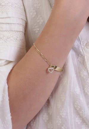 Gouden armband met een kettingontwerp en een hartvormige hanger met witte kristallen, gedragen op een lichtgekleurde arm tegen een witte textuurstof.