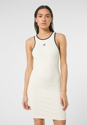 Frau trägt ein eng anliegendes, ärmelloses weißes Kleid mit schwarzem Rand und kleinem Logo auf der Brust, steht vor einem schlichten hellen Hintergrund.