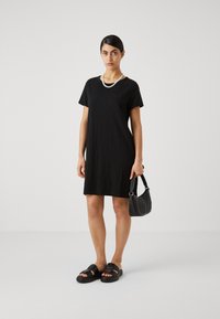 Svart t-skjortekjole med korte ermer, rund hals og en lomme. Kombinert med svarte sandaler og en liten svart håndveske.