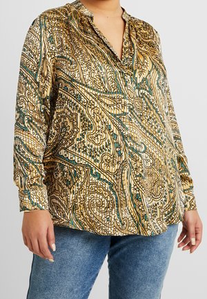 Nainen pukeutuneena pitkähihainen napilliseen paitaan, jossa on beigeitä, petroolinvihreitä ja ruskeita abstrakteja paisleymalleja, yhdistettynä sinisiin farkkuihin.