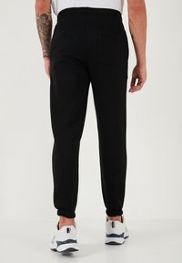 Crne trenirke s elastičnom waistband-om i manžetama na gležnjevima, s jednim stražnjim džepom i mekim tkaninom. Nosive s bijelim tenisicama.