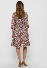 Mujer de espaldas, vestida con un vestido floral hasta la rodilla, con mangas abullonadas y zapatos de tacón altos marrones, contra un fondo liso.
