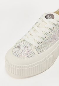 Bela superga z debelim podplatom, prekrita z majhnimi iridescentnimi kamenčki. Ima ravne vezalke in zaobljen sprednji del. Teksturiran gumijast podplat.