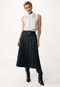 Gonna in pelle plissettata nera abbinata a una camicia bianca con dettaglio a fiocco. Il modello indossa stivali al ginocchio in pelle nera.