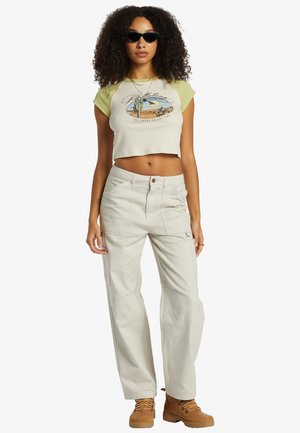 T-shirt vert clair et crème coupé avec un graphisme de désert, associé à un pantalon cargo beige et des bottes beige. Tissu lisse avec une coupe décontractée.