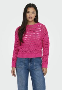 Roze gebreide trui met een open ruitpatroon, geribbelde ronde halslijn en manchetten, gecombineerd met hoog uitgesneden spijkerbroek.