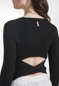 Schwarzes, geripptes Crop-Top mit langen Ärmeln, das einen ausgeschnittenen Rücken und ein überkreuztes Design aufweist. Etikett am Halsausschnitt in Hellrosa sichtbar.