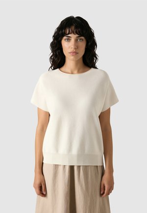 Žena s tmavými kudrnatými vlasy, oblečená v krémově bílém krátkém rukávu a béžové sukni, stojící před jednobarevným světle šedým pozadím.