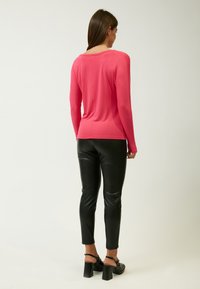 Rosa Langarmoberteil aus glattem Stoff, kombiniert mit enganliegenden schwarzen Kunstlederhosen. Die Rückansicht zeigt einen runden Halsausschnitt und eine schmale Passform.