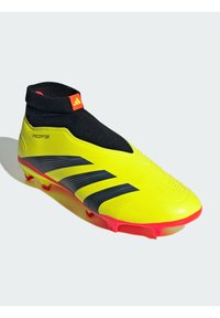 Botín de fútbol amarillo neón con un collar de tobillo negro tipo calcetín, tres franjas negras y suela roja. Presenta un upper texturizado y un diseño elegante.