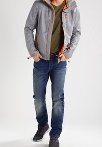 Grijze capuchonjas met oranje accenten over een groen T-shirt, gecombineerd met blauwe spijkerbroek en grijze sneakers. Voorzien van ritsen en zakken.