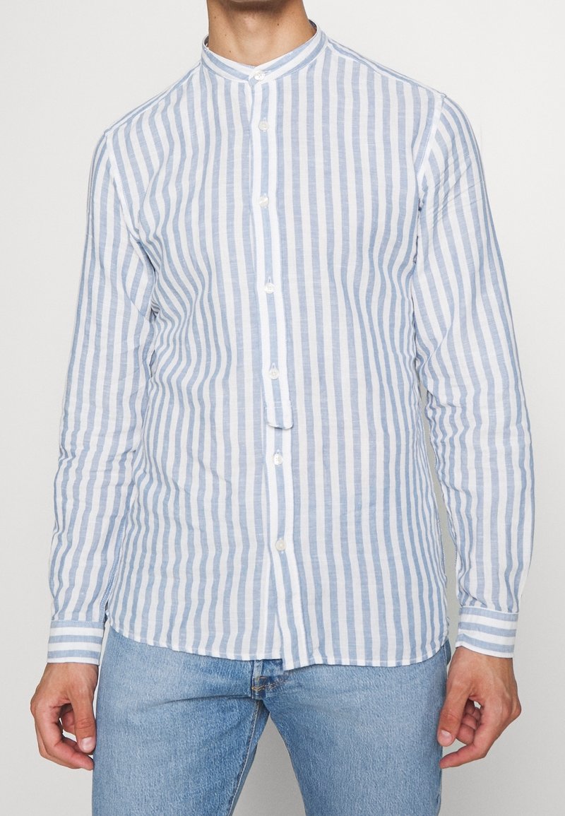 Homme portant une chemise à boutons à manches longues avec des rayures verticales blanches et bleu clair, col mao, et un jean bleu clair.