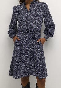 Abito nero fiorato con motivi viola e verdi, maniche lunghe a sbuffo, chiusura frontale a bottoni, cintura regolabile in vita e tasche laterali.