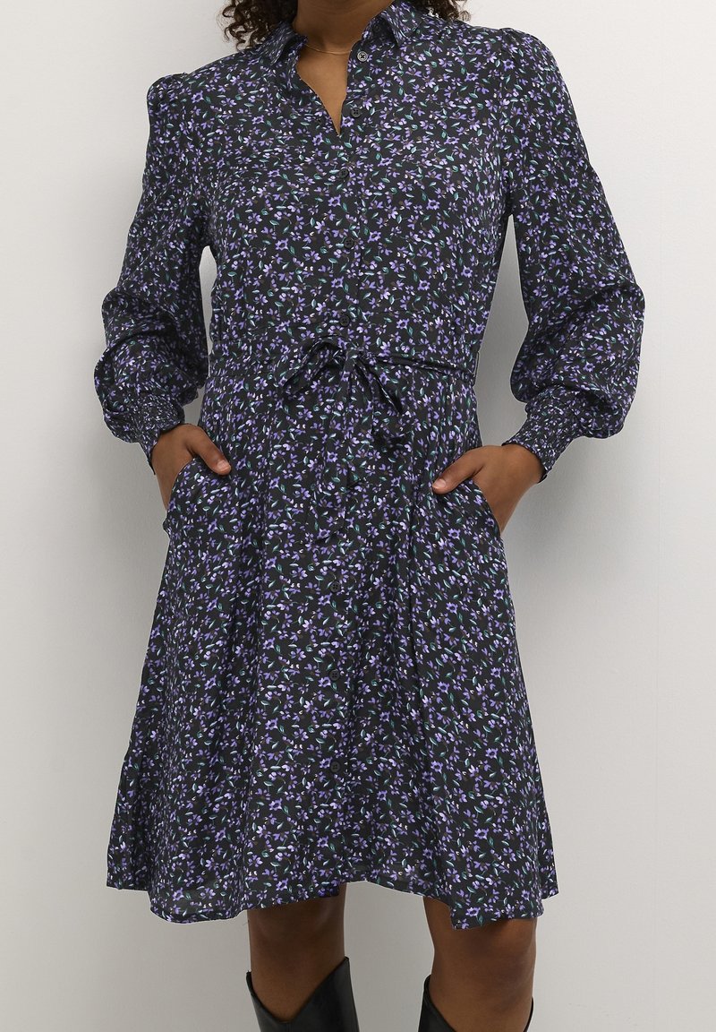 Abito nero fiorato con motivi viola e verdi, maniche lunghe a sbuffo, chiusura frontale a bottoni, cintura regolabile in vita e tasche laterali.