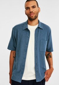 Kurzarm-Hemd mit Knopfleiste aus blauem Strukt Gewebe, mit Kragen, Brusttasche und runden Knöpfen, über einem weißen T-Shirt getragen.