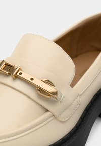 Mocassin en cuir crème avec détail de boucle métal dorée et semelle en caoutchouc noire, montrant un gros plan des coutures avant et latérales.