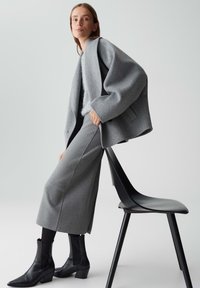 Grauer Wollmantel mit weiten Ärmeln, kombiniert mit einem hellgrauen Strickpullover und passendem Rock. Schwarze Stiefeletten runden das Outfit ab.