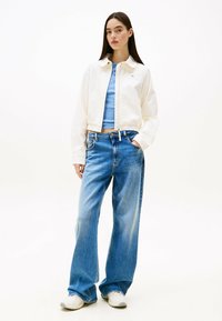 Witte cropped jas met een ronde kraag over een lichtblauwe getailleerde top, gecombineerd met losse lichtblauwe jeans en witte sneakers.