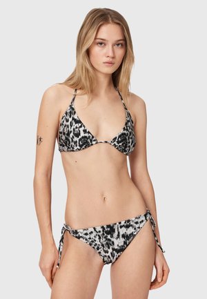 Ung kvinde iført en sort og hvid leopardprint bikini med siderem, står mod en ensfarvet lysegrå baggrund.