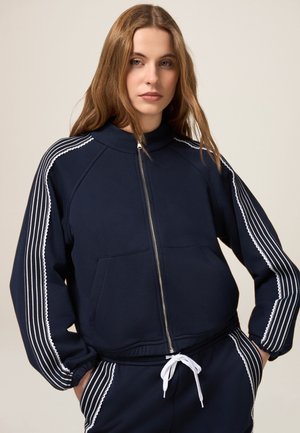 Navy zip-up sweatshirt with a high collar, white striped accents along sleeves, and a front pocket. Features a textured fabric and fitted design.