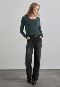 Ciemnozielony sweter z szerokim dekoltem i prążkowanymi mankietami, zestawiony z luźnymi czarnymi dżinsami i czarnymi botkami na obcasie w kształcie grota.