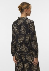 Vrouw met blond haar in een knot, gekleed in een zwart shirt met lange mouwen en een broek met beige bladpatronen, gezien van achteren tegen een witte achtergrond.