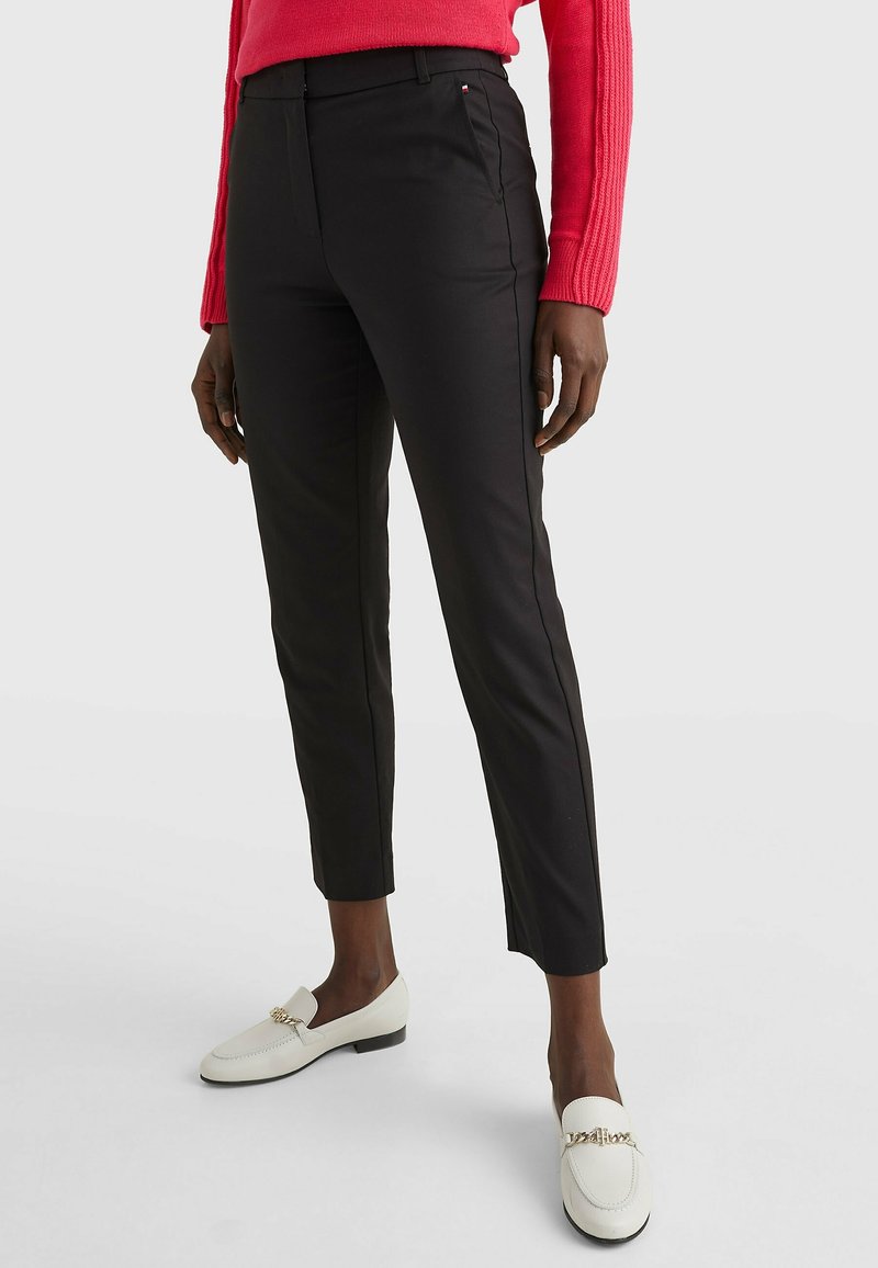 Pantalon noir sur mesure à coupe slim, avec une texture lisse et des détails latéraux subtils. Associé à des mocassins blancs.