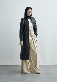 Black leather trench coat, beige sweater vest with double buttons, white shirt, and wide ivory trousers. Smooth textures and layered design.