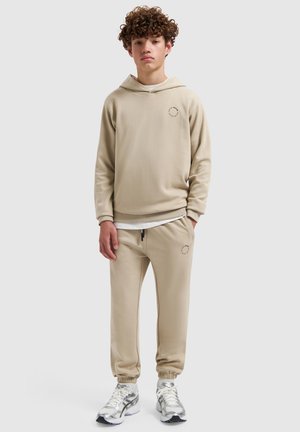 Teenager mit lockigem Haar, der einen beigen Hoodie und Jogginghosen sowie weiße Turnschuhe vor einem schlichten Hintergrund trägt.