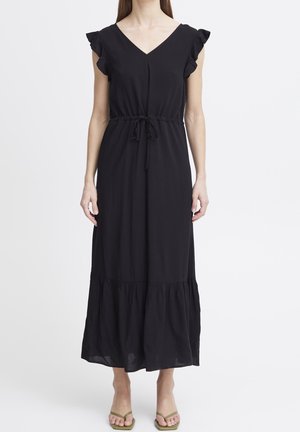 Kobieta ubrana w czarną, bezrękawnikową suknię maxi z falbanami na ramionach, z paskiem do ściągania w pasie i falbanami na dole, zestawioną z zielonymi sandałami.