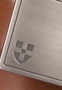 Szczotkowana metalowa płyta z wygrawerowanym logo przedstawiającym tarczę i wzór siatki, umieszczona na fakturowanej brązowej skórze.