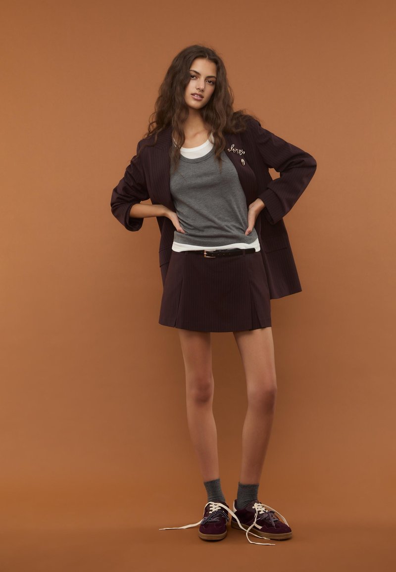 Blazer à rayures noires et violettes, t-shirt gris superposé sur un débardeur blanc, jupe courte à rayures, chaussettes grises, chaussures bordeaux avec lacets blancs.