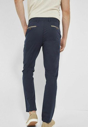 Mann trägt navyblaue, schmal geschnittene Hose mit geknöpften Rückentaschen und beigefarbenen Slip-on-Schuhen, steht vor einem einfarbigen Hintergrund.