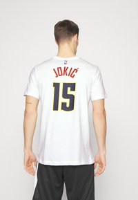 T-shirt blanche à manches courtes avec le nom "JOKIĆ" et le numéro "15" en rouge et bleu marine, avec une étiquette du logo NBA au niveau de l'encolure.