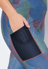 Leggings de colores y patrones en azul, verde, rojo y blanco, con un bolsillo de malla negra y detalles de costura en contraste. Textura elástica y suave.