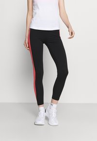 Person som bär svarta leggings med röda sidstripe, en vit linne, vita Puma-strumpor och vita sneakers, stående mot en neutral bakgrund.