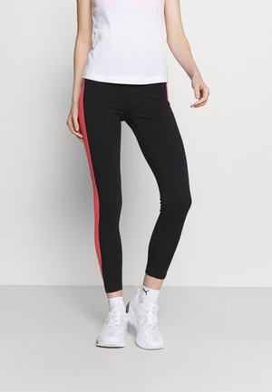 Person wearing black leggings with red side stripes, a white tank top, white Puma socks, and white sneakers, standing against a plain background.