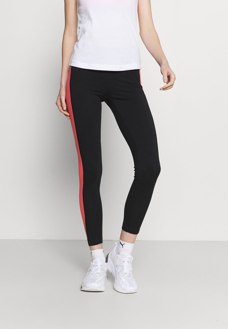 Person som bär svarta leggings med röda sidstripe, en vit linne, vita Puma-strumpor och vita sneakers, stående mot en neutral bakgrund.