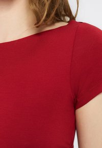 Κοντινό πλάνο ενός ατόμου που φοράει εφαρμοστό, κόκκινο πλεκτό τοπ με κοντά μανίκια και φαρδύ ντεκολτέ, που φαίνεται ο ώμος και μέρος του λαιμού.