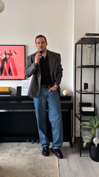 Vyras vilki pilką dryžuotą švarką ant juodo polo marškinėlio, mėlynas džinsus ir juodus batus. Jis stovi šalia pianino ir juodo metalinio lentynos su dekoratyviniais daiktais.