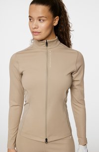 Beige zip-up idrottsjacka med långa ärmar, texturerad tyg och en smal passform. Har en hög krage och logotyp tryckt fram.