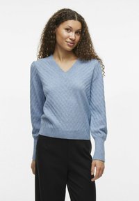 Hellblauer, langärmeliger Pullover mit V-Ausschnitt, der ein strukturiertes diagonales Muster und einen gerippten Saum aufweist, kombiniert mit schwarzen Hosen.