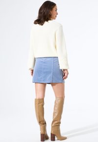 Frau, die einen cremefarbenen Strickpullover, einen hellblauen Cordminirock und braune kniehohe Lederstiefel trägt und mit dem Rücken zur Kamera steht.