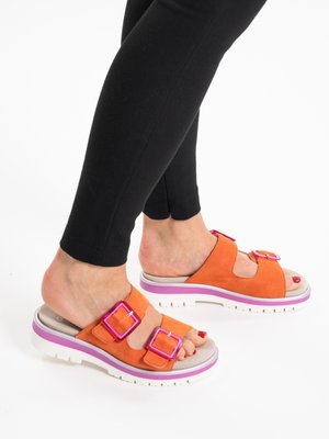 Pieds en sandales à glissière orange avec boucles roses et semelles blanches, portant des leggings noirs sur fond blanc.