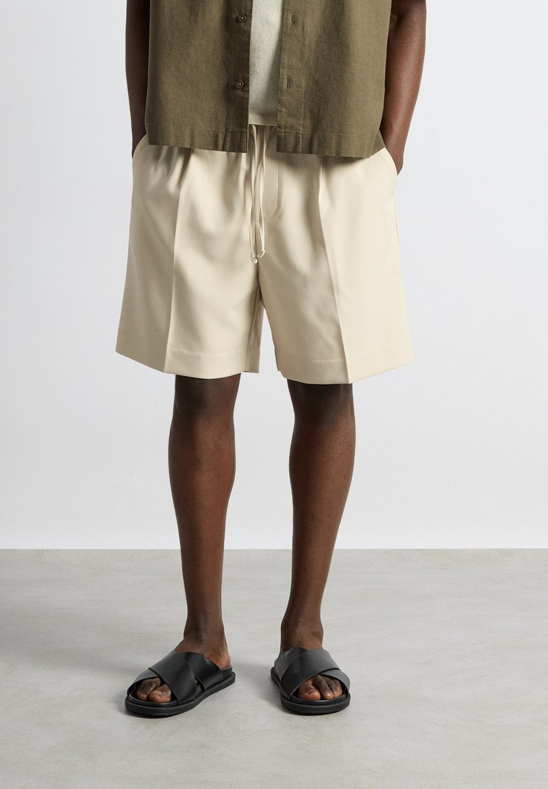 Personne portant un short beige à cordon, une chemise à manches courtes vert olive et des sandales noires croisées, debout sur un sol gris.