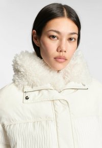 Vadderad vit jacka med en fluffig, texturerad krage, som har dragkedjestängning och plisserad design på kroppen. Slät innersida.